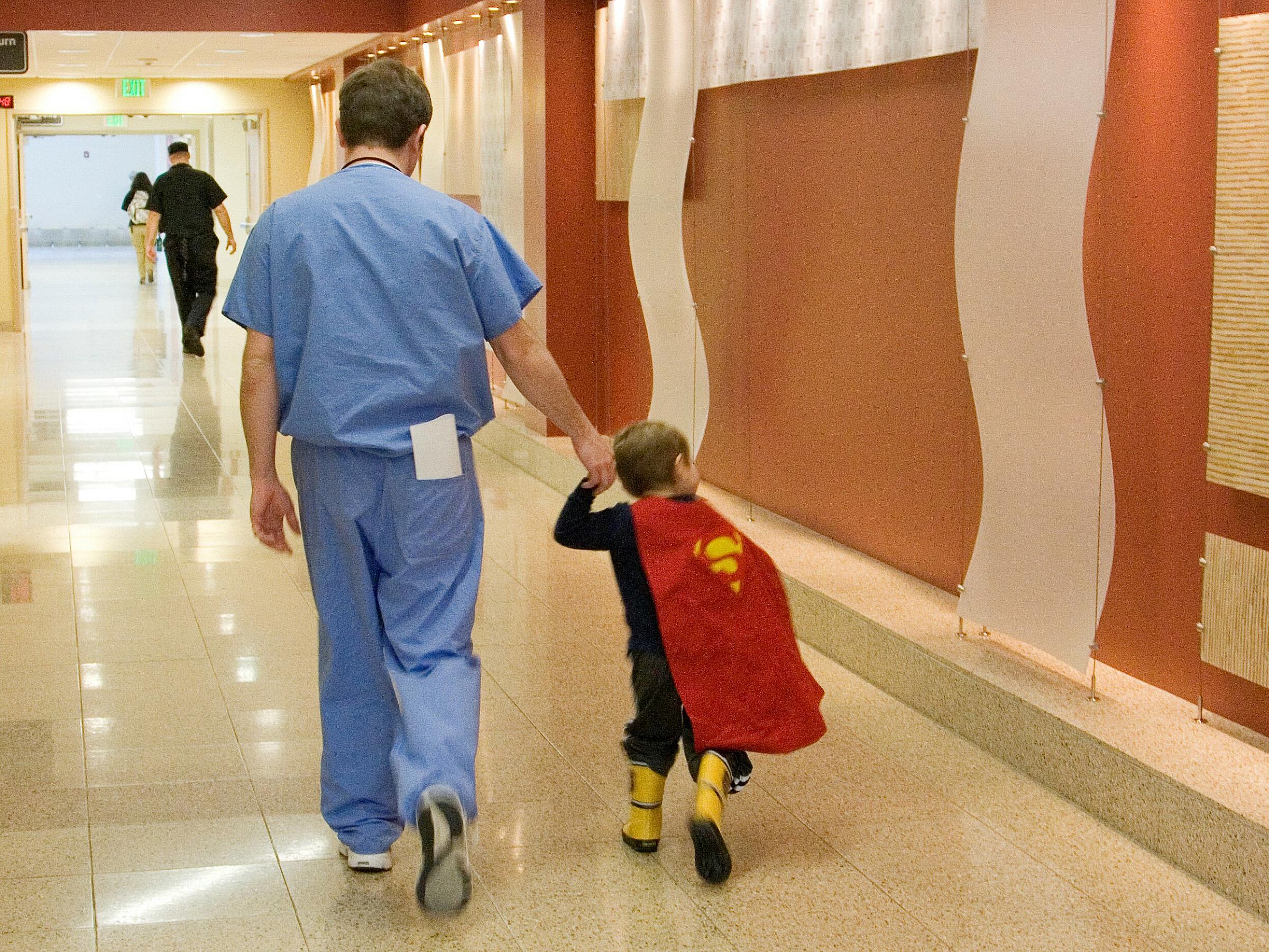 Brock O'Neil wearing scrubs as he walks down the hallway at Huntsman Cancer Institute while holding his son's hand who is wearing a superhero cape