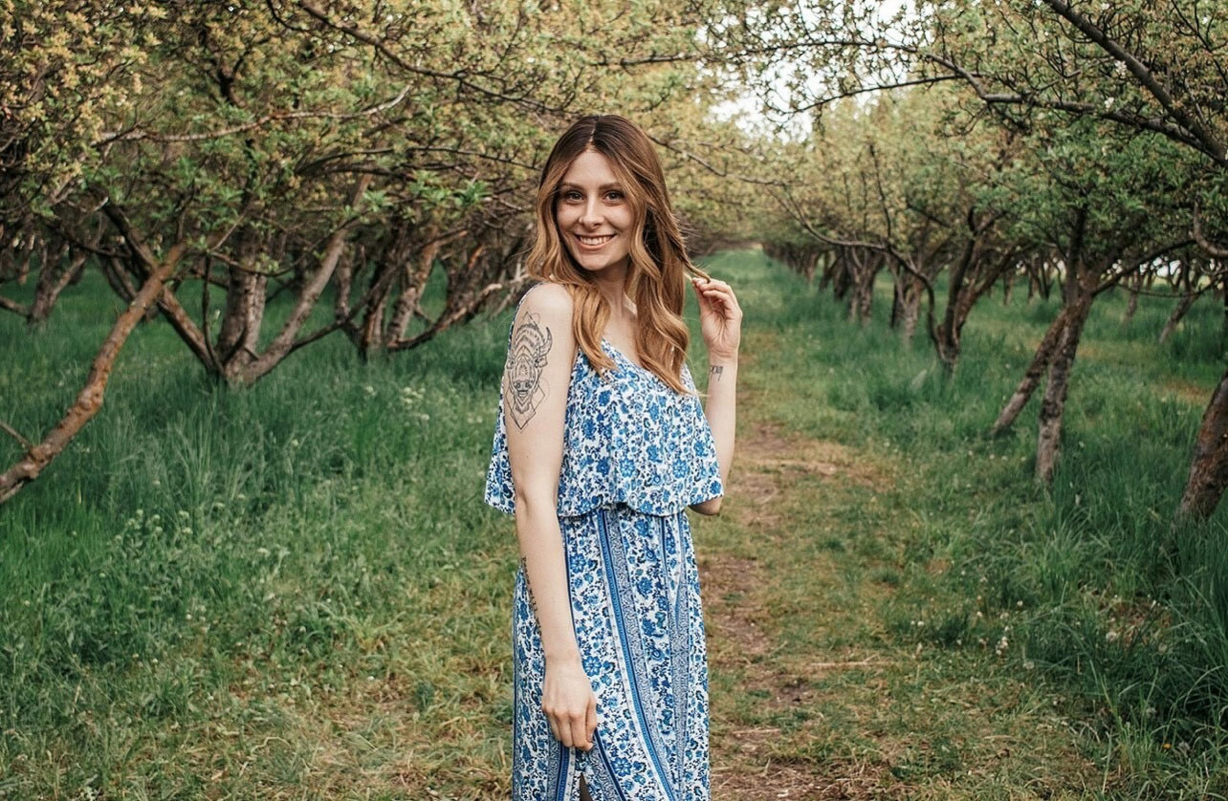 Tylar Bonnell standing between rows in an orchard in a sundress and playing with her hair