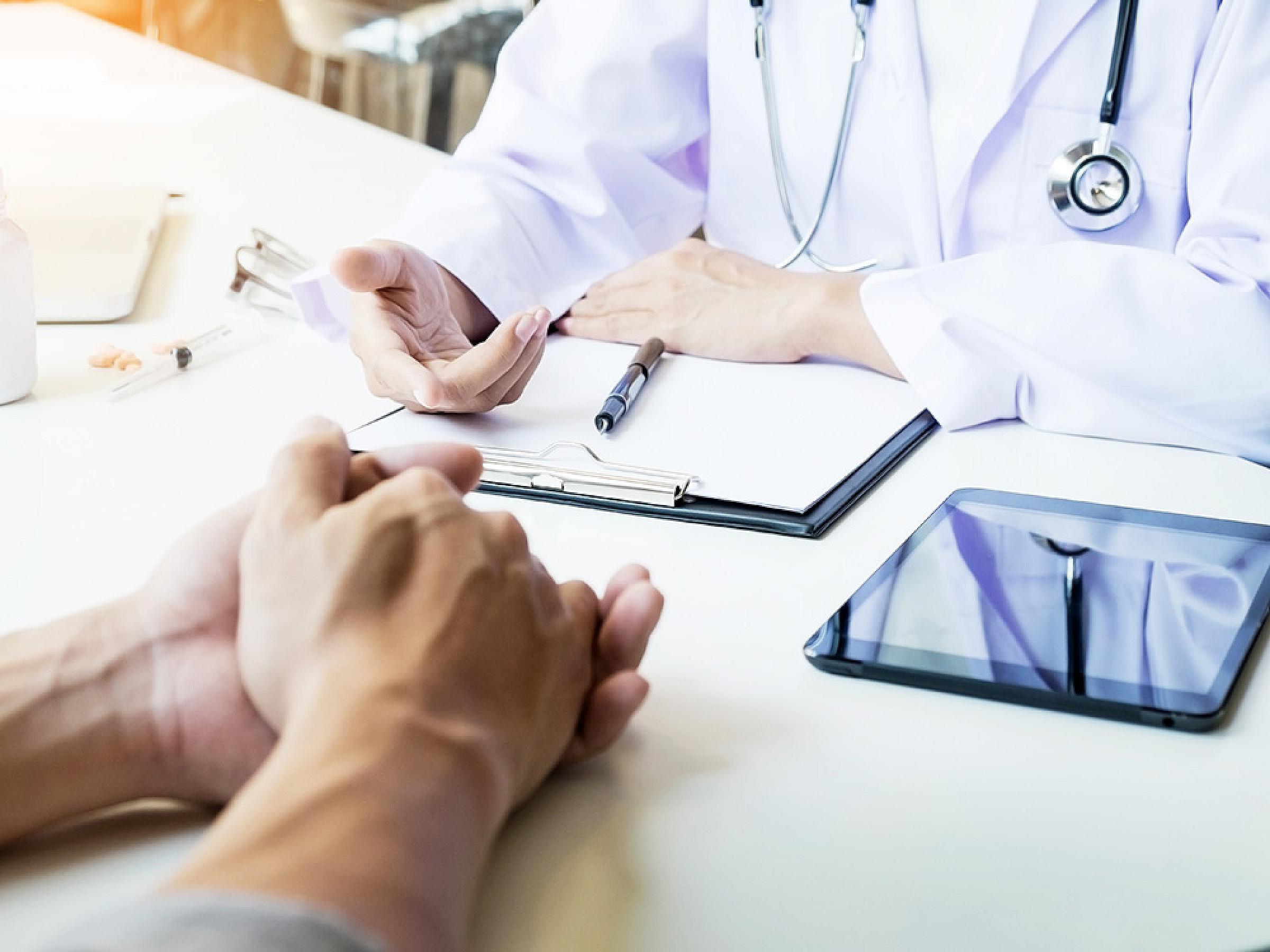 Doctor Discussing with Visitor Over Desk with Clipboard and Tablet