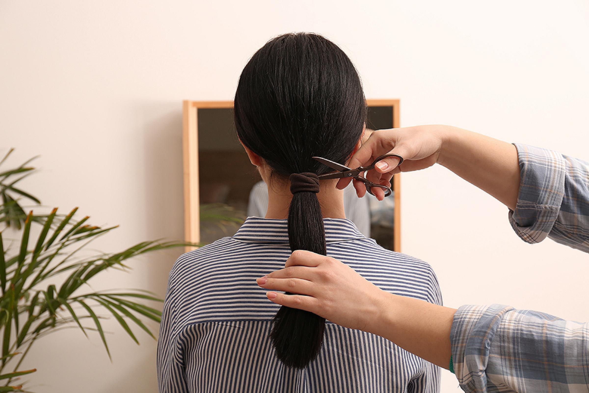 Woman facing away from camera while another person prepares to cut her ponytail off