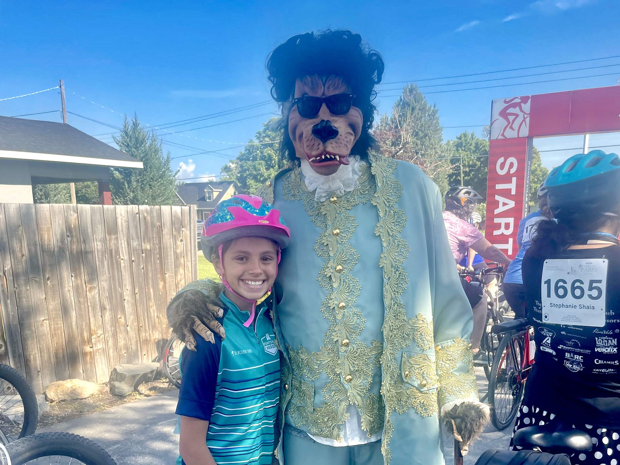 Person wearing ornate clothing and a monster dog mask with a curly wig on top poses with a young Little Red participant