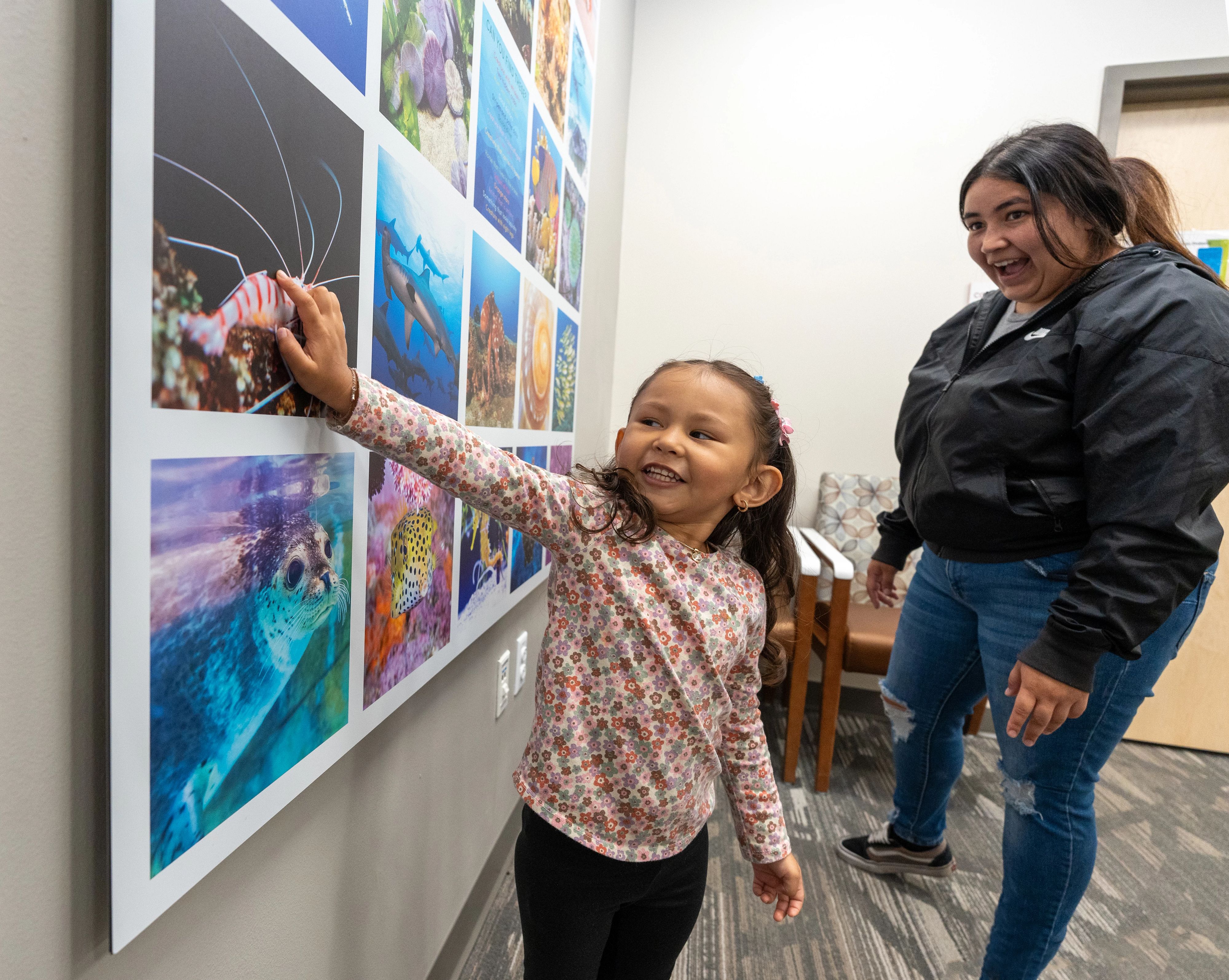 Little girl with tan skin and pigtails smiles and touches aquatic pictures on the wall. 