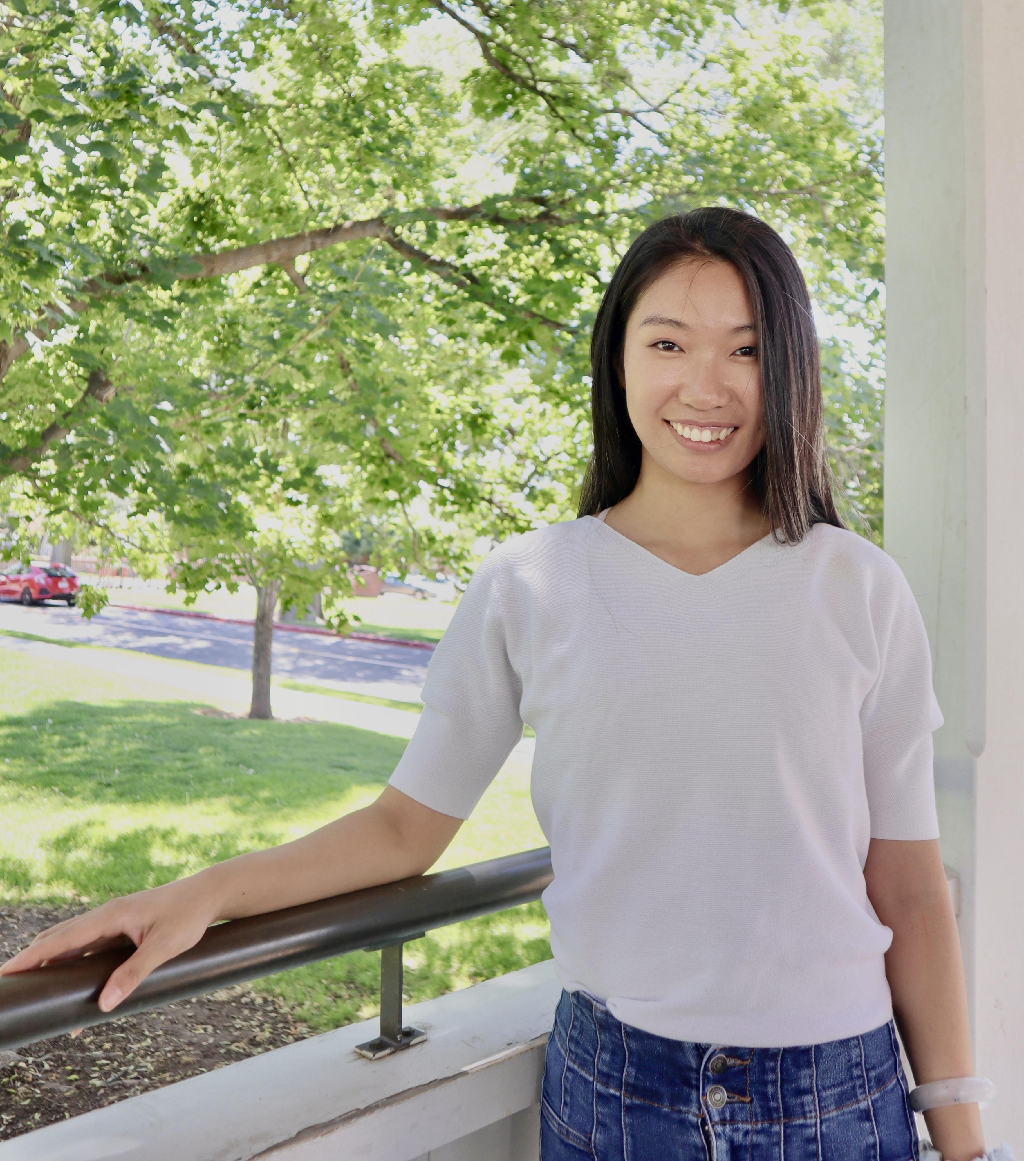A person in a white shirt smiles in front of a sunlit tree.