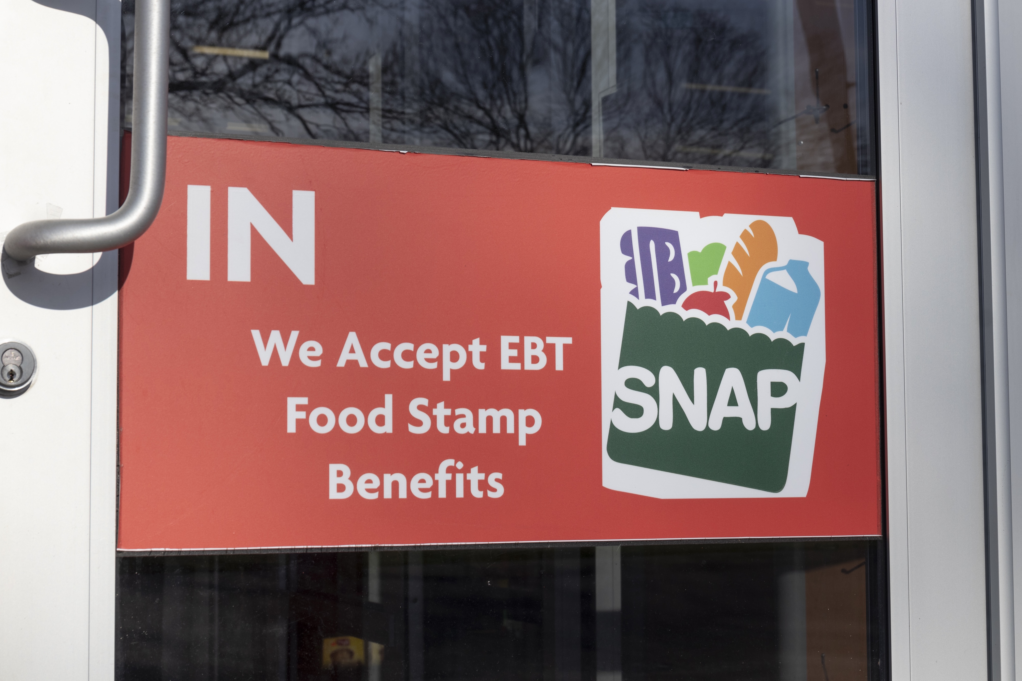 Photo of a doorfront sign reading "IN. We accept EBT food stamp benefits" next to the SNAP logo of a full grocery bag.