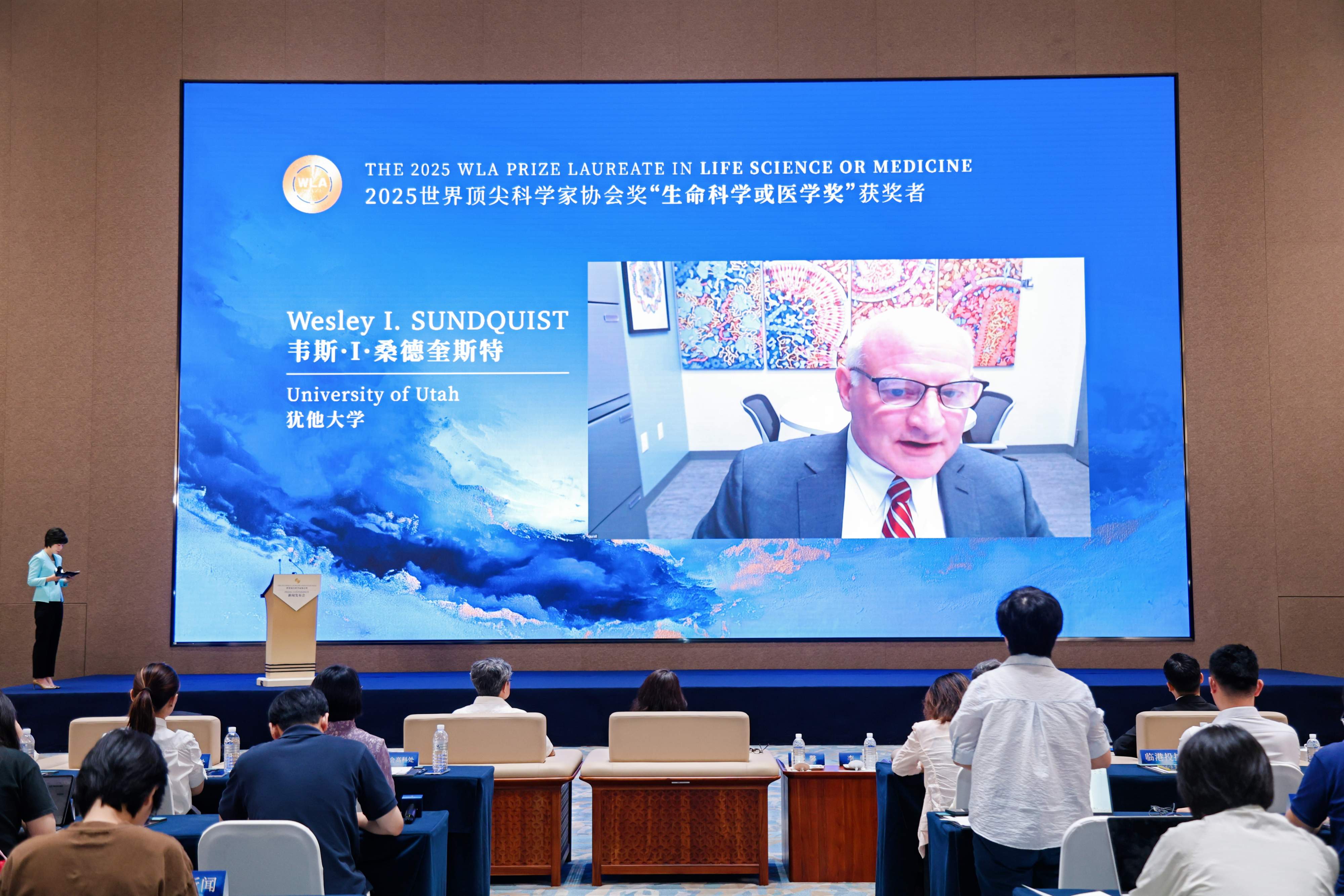 Photo of a powerpoint slide showing a livestream of a man in a suit on a fancy blue background.