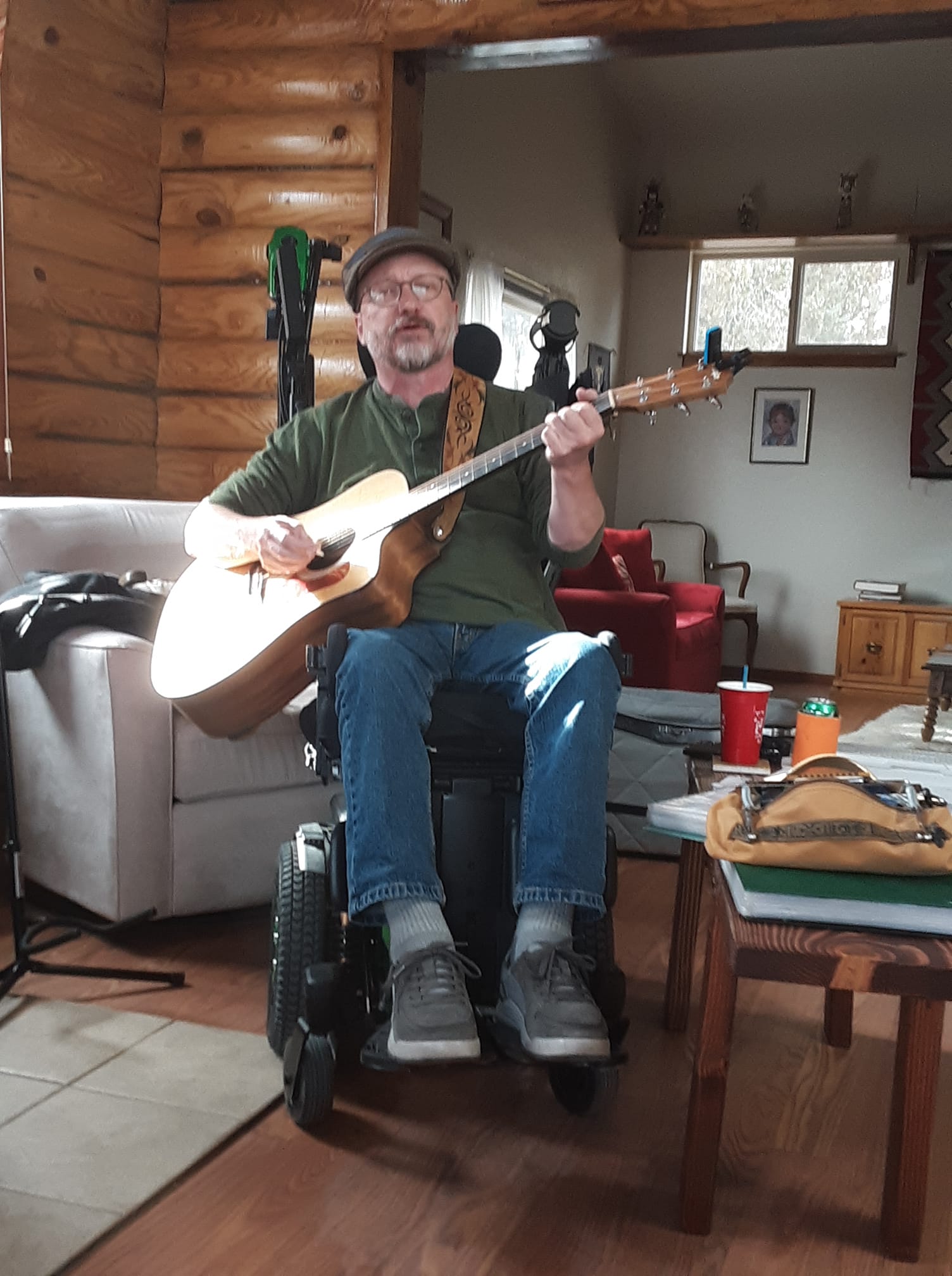 A person wearing a green shirt and flat cap strums a guitar and sings. He's sitting in a power wheelchair inside a log cabin.
