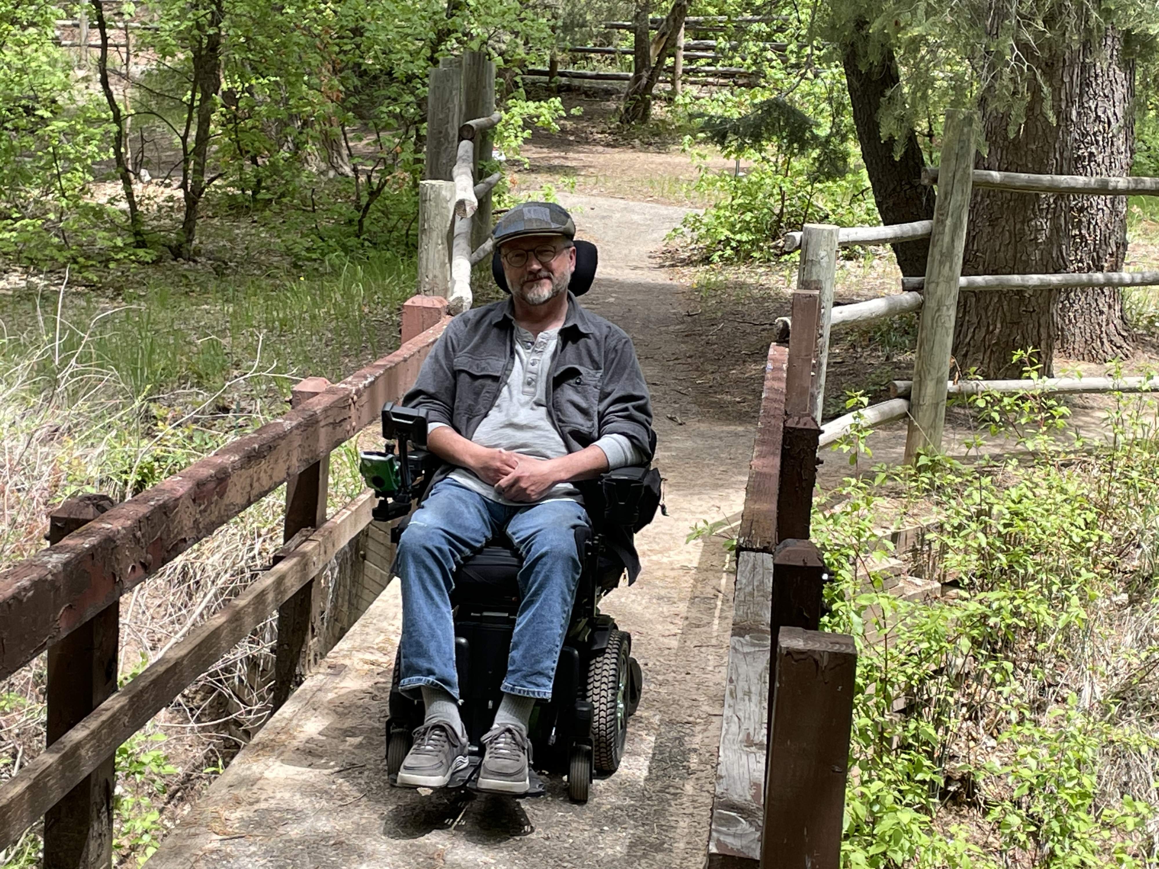 A person in a black jacket and flat cap smiles at the camera. He's in a power wheelchair on a bridge in the woods.