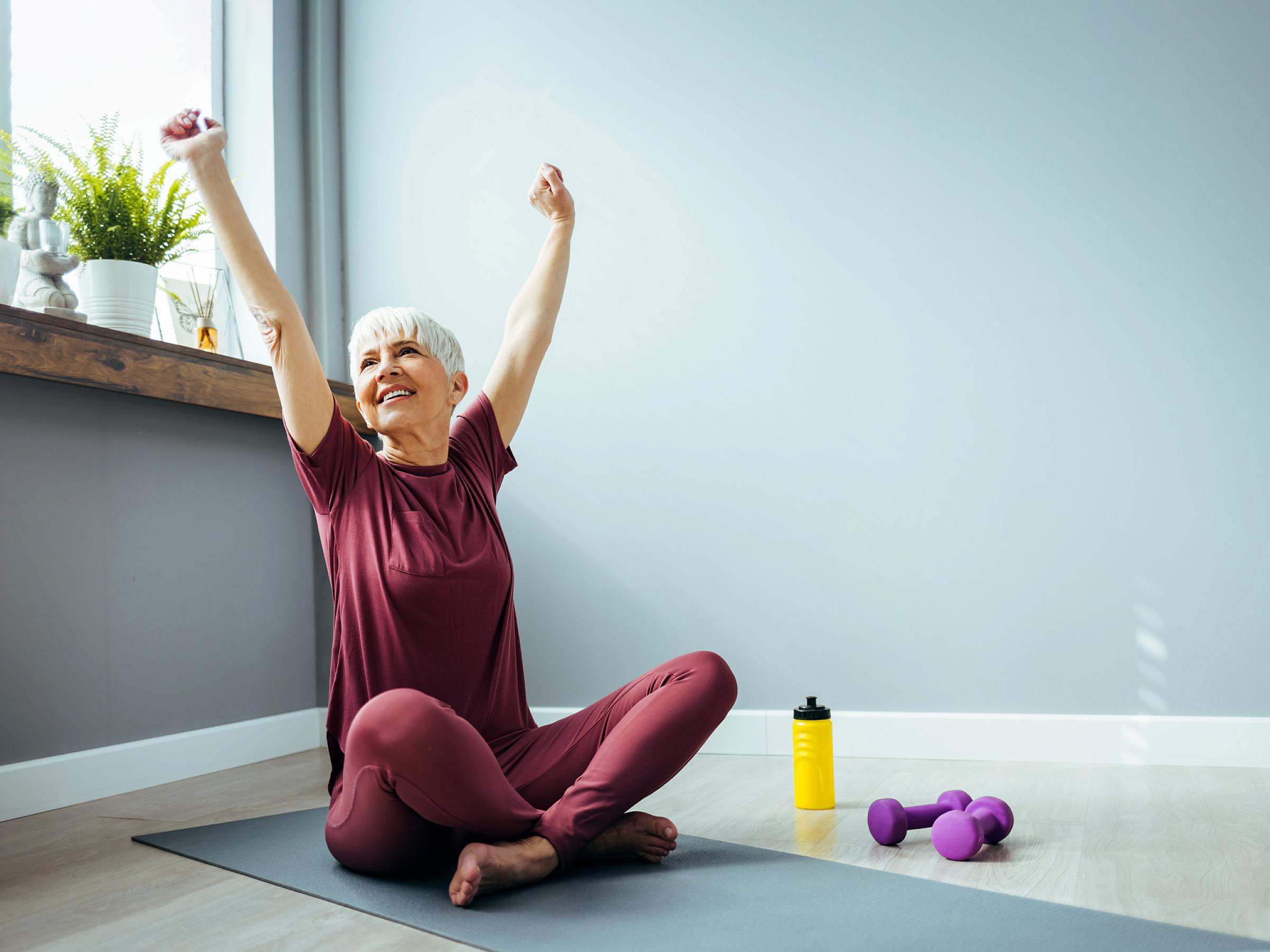 Older woman sits on a yoga mat and stretches her arms in the air