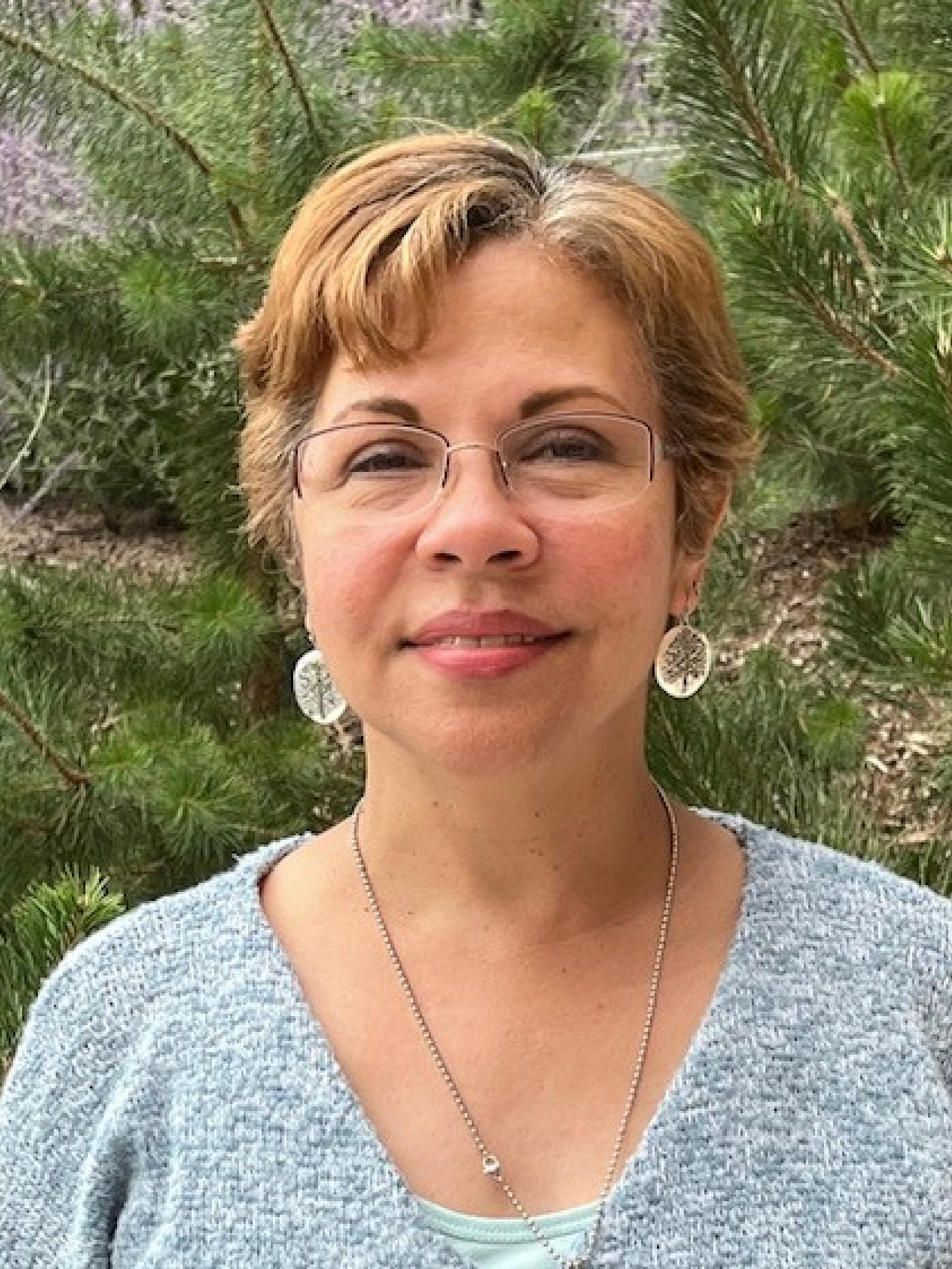 Woman with short blonde hair smiles by a pine tree