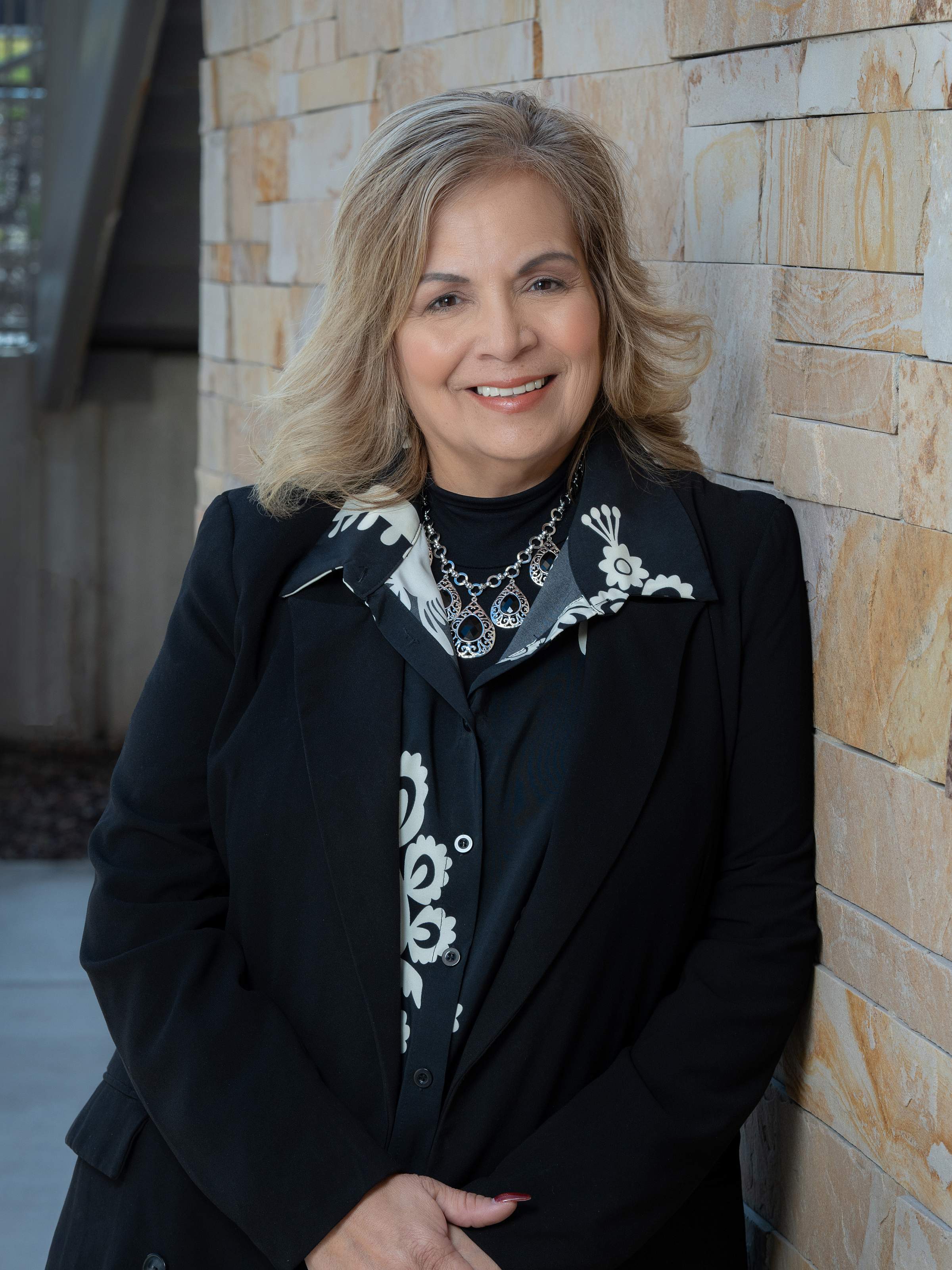 A woman with medium-length blonde hair wears a black coat and poses for a professional headshot