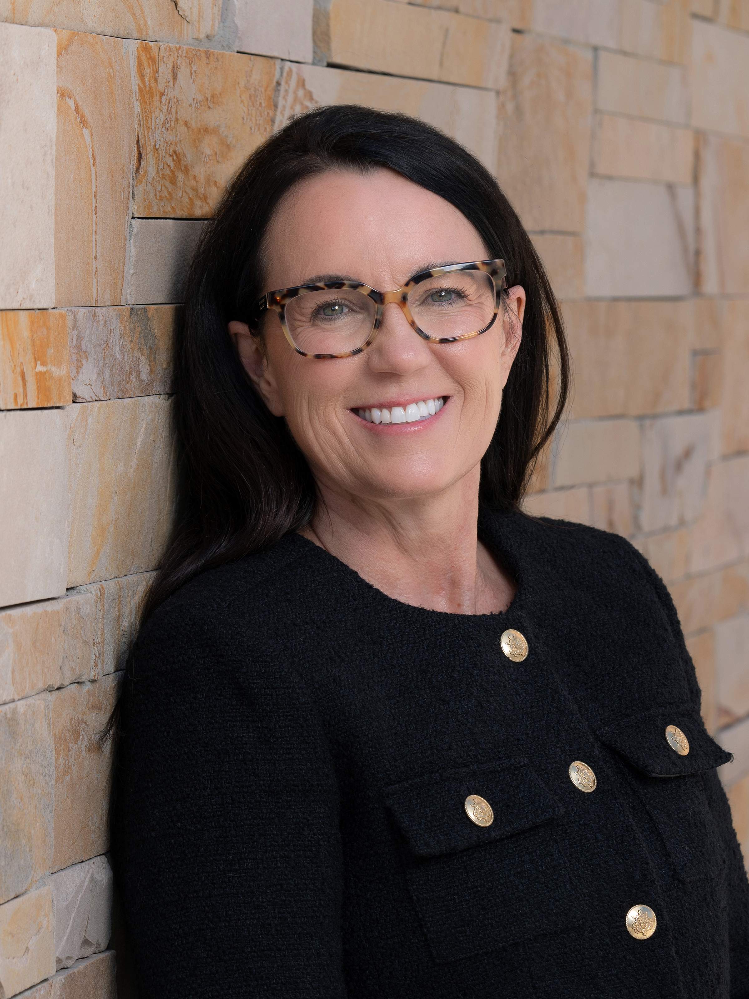 A woman with long black hair and glasses wears a black jacket and poses for a professional headshot