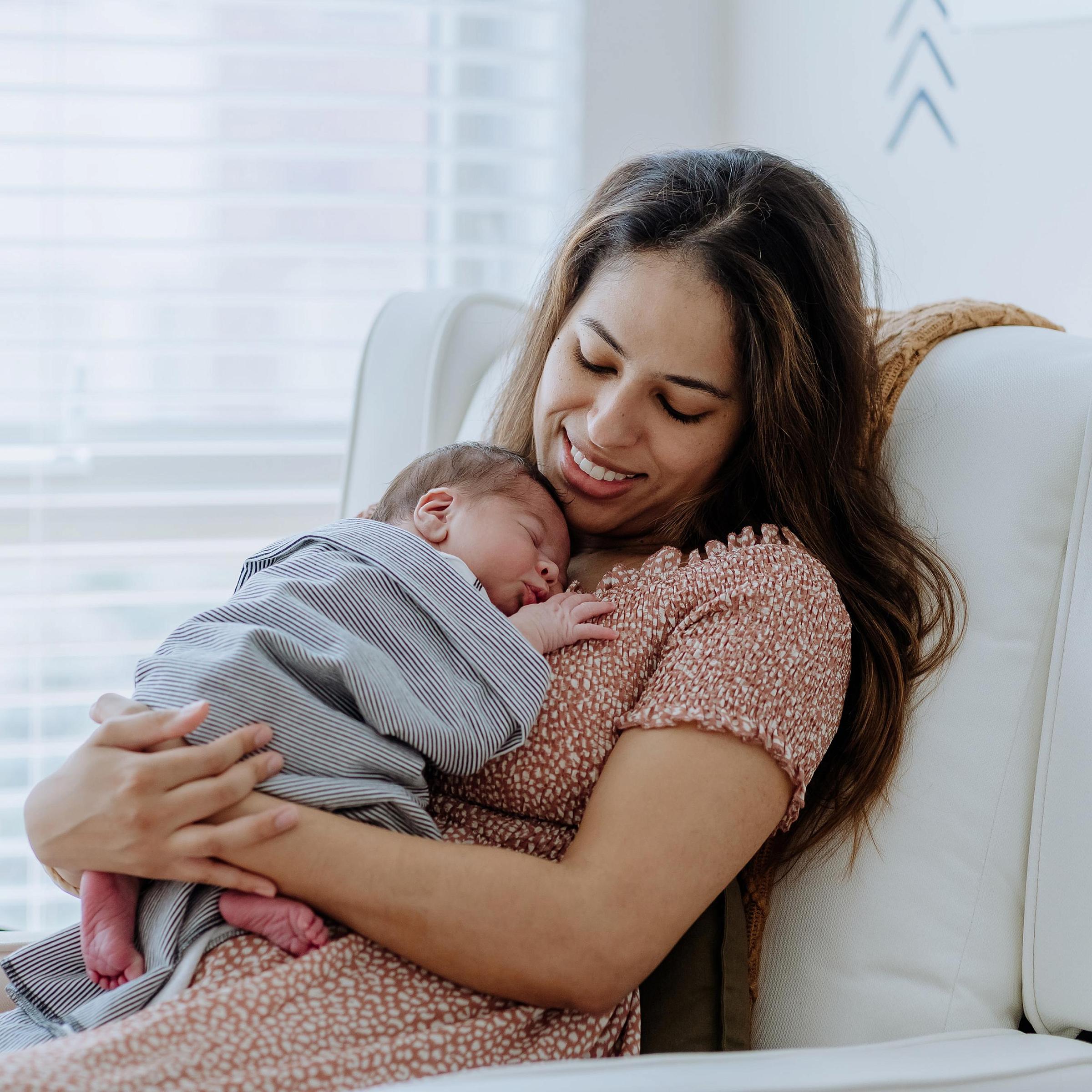 Mother with long dark hair in a floral dress holds her baby in chair