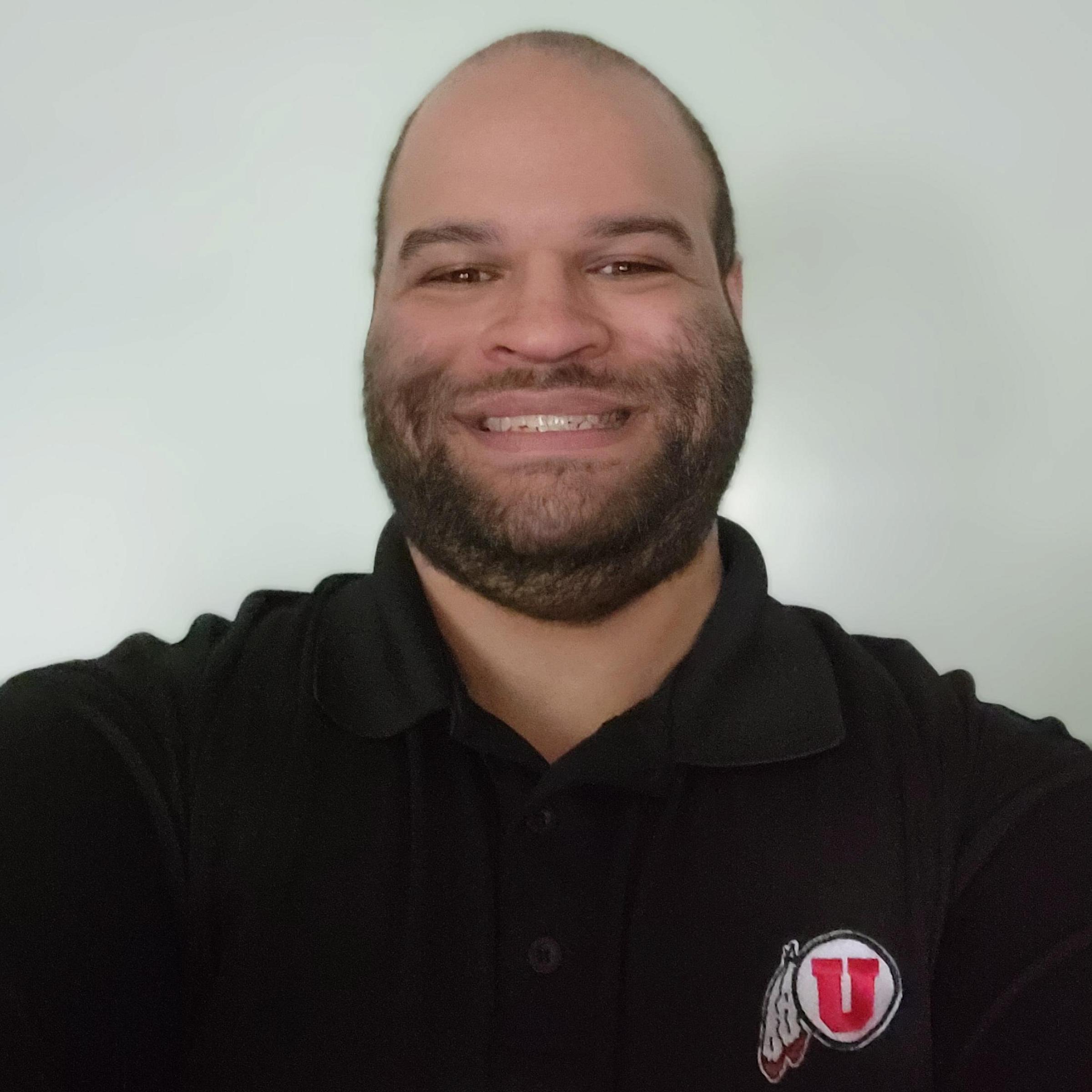 Matthew Tydeck has a shaved head, facial hair, and wears a black polo with the University of Utah logo.