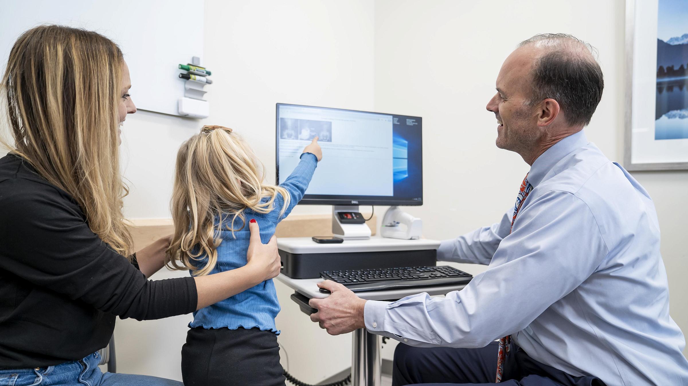 A little girl sits on her mom's lap and points at a computer next to a U of U Health urologist